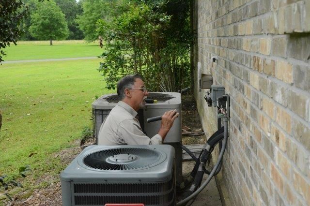 Man inspects an AC unit beside a brick wall. Green grass and trees are in the background.