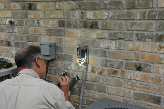 Man inspecting an outdoor electrical outlet on a brick wall, using a flashlight.