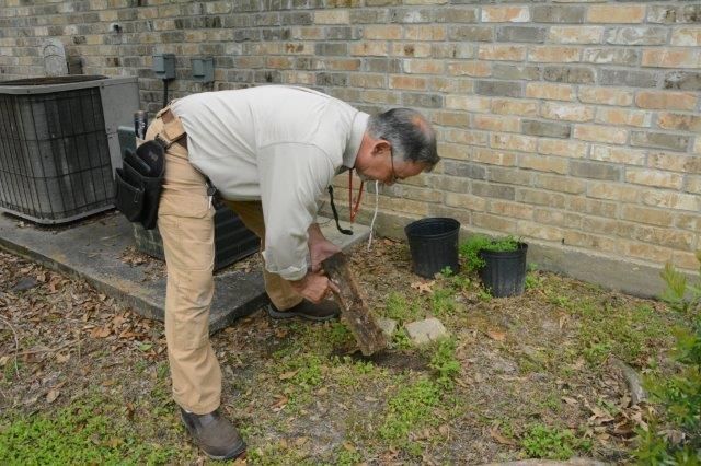 Man digging in yard near a brick building. He wears light clothing, bent over, focused. Black air conditioner visible.
