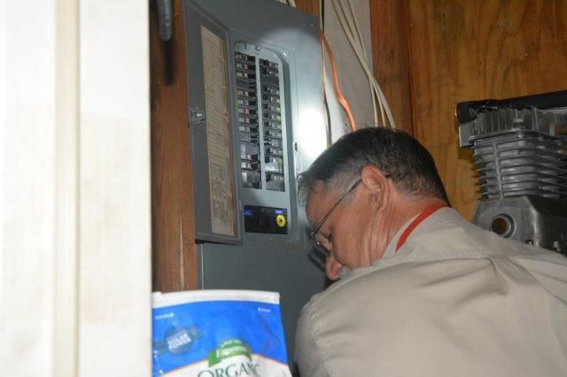 Man inspecting a breaker box. He's wearing glasses and a light-colored shirt. A blue bag is on the floor.
