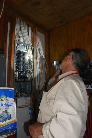 Man inspecting an electrical panel with exposed wires in a wood-paneled room.