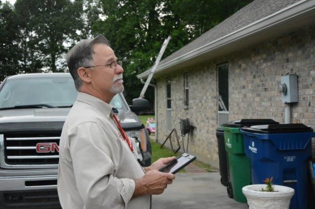 Man examining a house exterior with a tablet; silver truck and trash cans are visible.