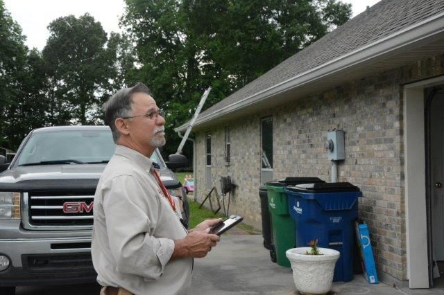 Man inspecting a house, holding a clipboard, looking up at the roof. Gray truck, green and blue trash cans are nearby.