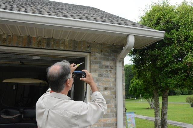 Man taking photo of a house gutter. Gray roof, brick wall, garage door, green tree.