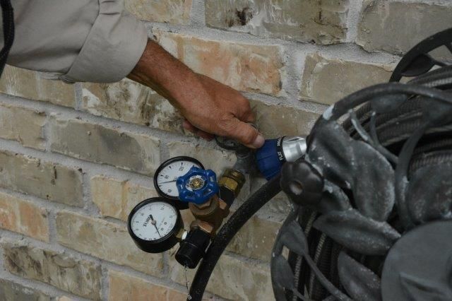 Person's hand adjusting a blue valve attached to a brick wall with gauges and a hose nearby.