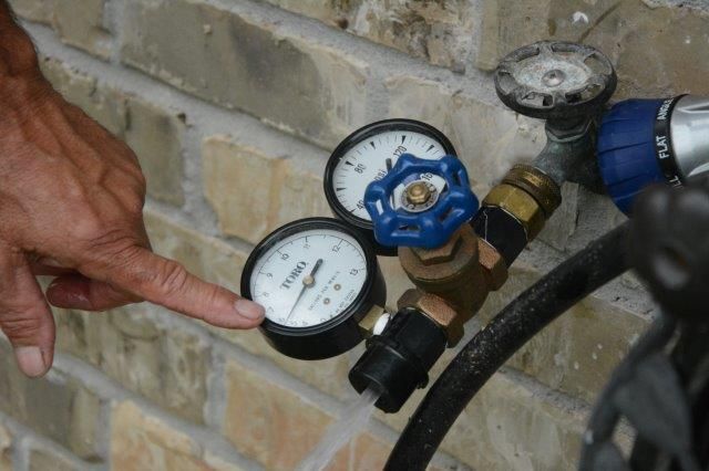 Hand pointing at a pressure gauge on an outdoor spigot with two gauges and a blue handle.