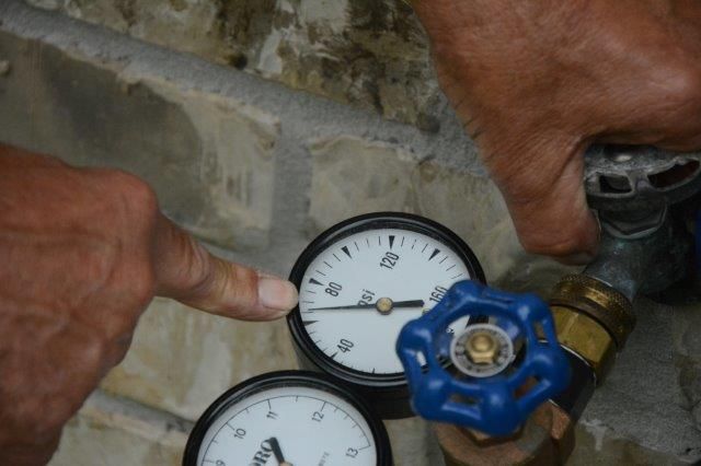 Hands pointing at a water pressure gauge reading approximately 60 psi, near a blue-handled valve.