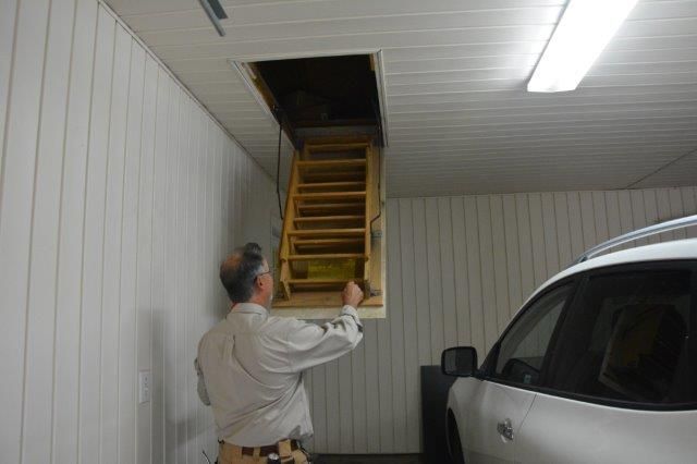 Man opens attic access door with pull-down stairs in a garage. A white car is parked nearby.