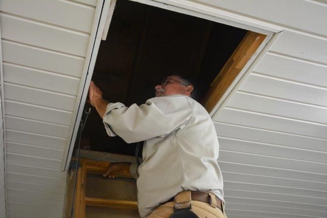 Man on ladder, reaching into attic opening in white ceiling.
