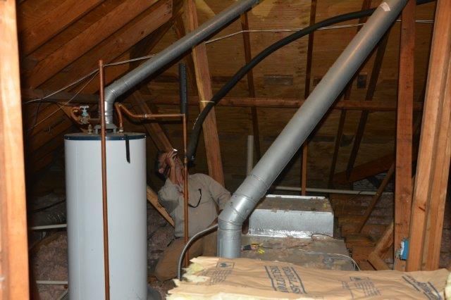 Person inspecting a water heater and furnace in an attic with wooden beams, insulation, and metal pipes.