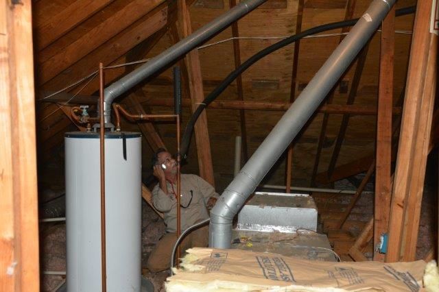Man examining water heater and flue pipes in a dusty attic with wood framing.