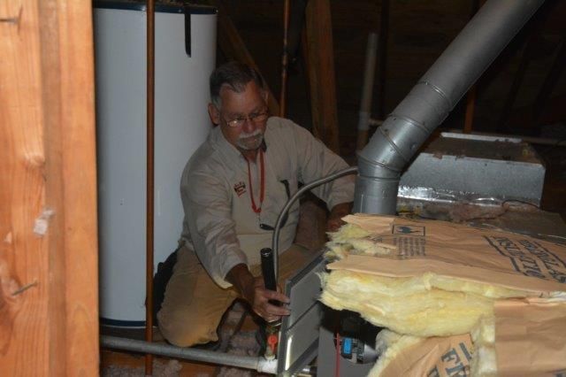 Man inspecting HVAC system in attic. Light brown clothing, insulation, metal ductwork.