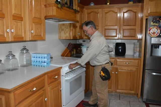 Man in kitchen adjusting stove, light wood cabinets, white countertops, black appliance.