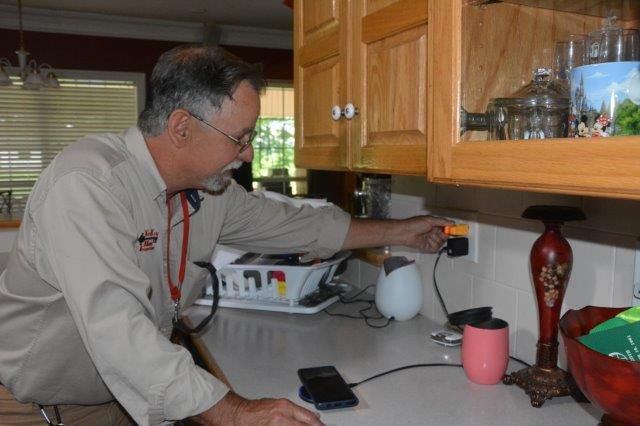 Man tests electrical outlet in a kitchen, smiling. Cabinets and countertop are visible.