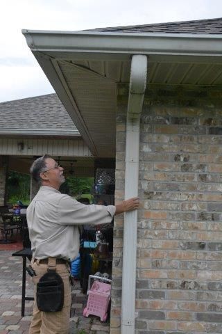 Man inspecting a white downspout on a brick wall next to a house's roof and gutter.