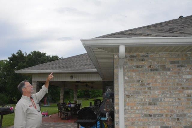 Man points upward at a house's roofline with gutters. Cloudy sky, brick wall, patio furniture visible.