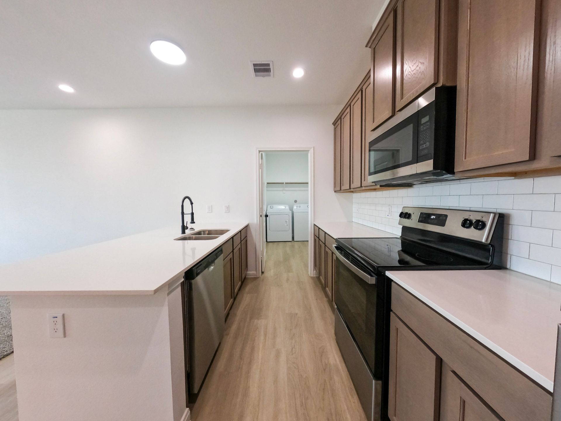 Modern kitchen with island, stainless steel appliances, and laundry room visible. Light wood cabinets and floors.