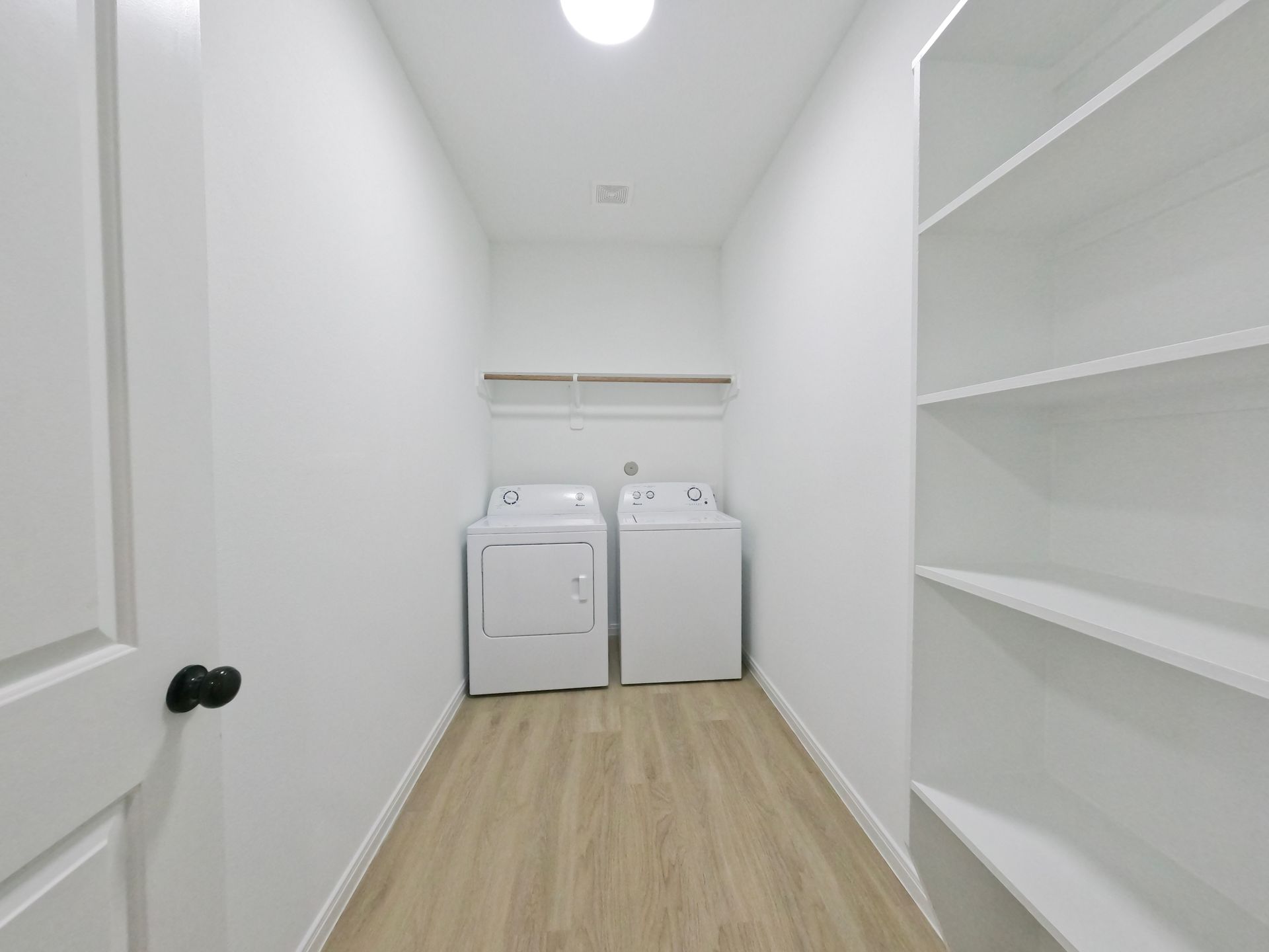 Laundry room with washer, dryer, and shelves. White walls, wood-look floor, and a closed door.