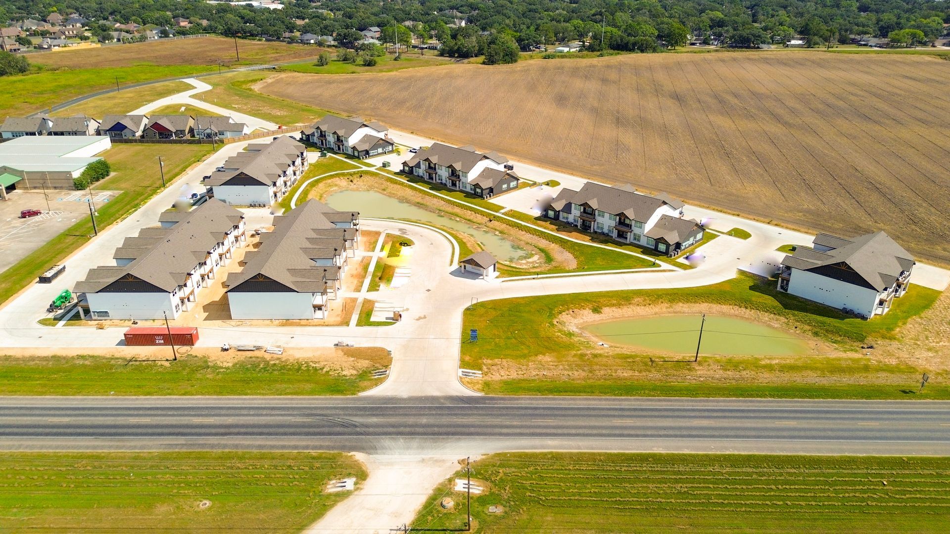 Aerial view of townhomes with a road and a field in the background. Green grass and a small pond are in the foreground.