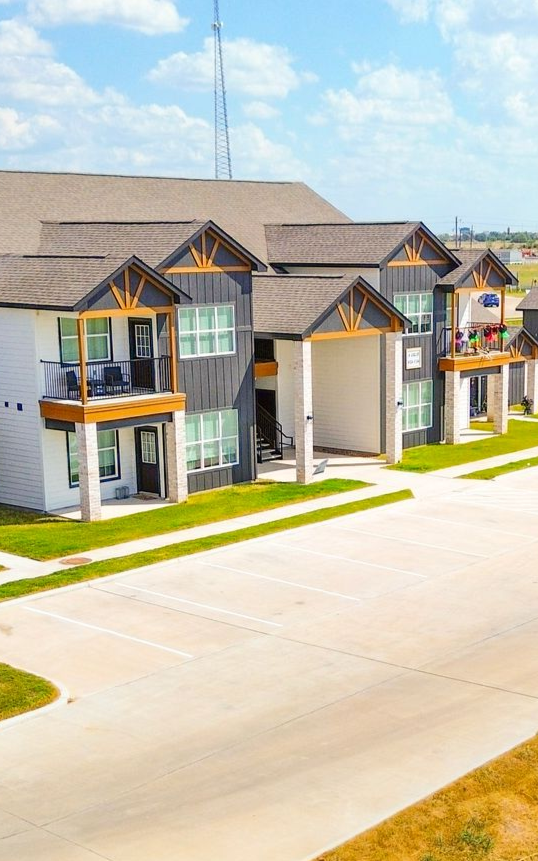 Apartment complex with gray and white siding, balconies, and brown roofs, on a sunny day.