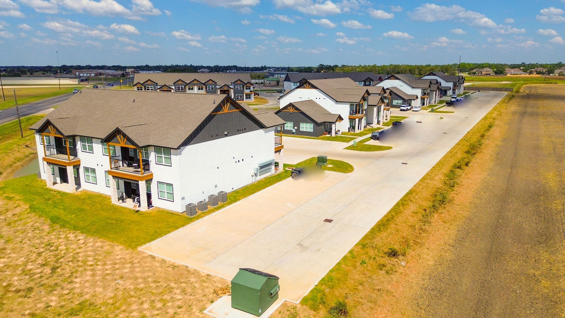Exterior view of modern white apartments with brown roofs and parking lot, set in a rural area on a sunny day.