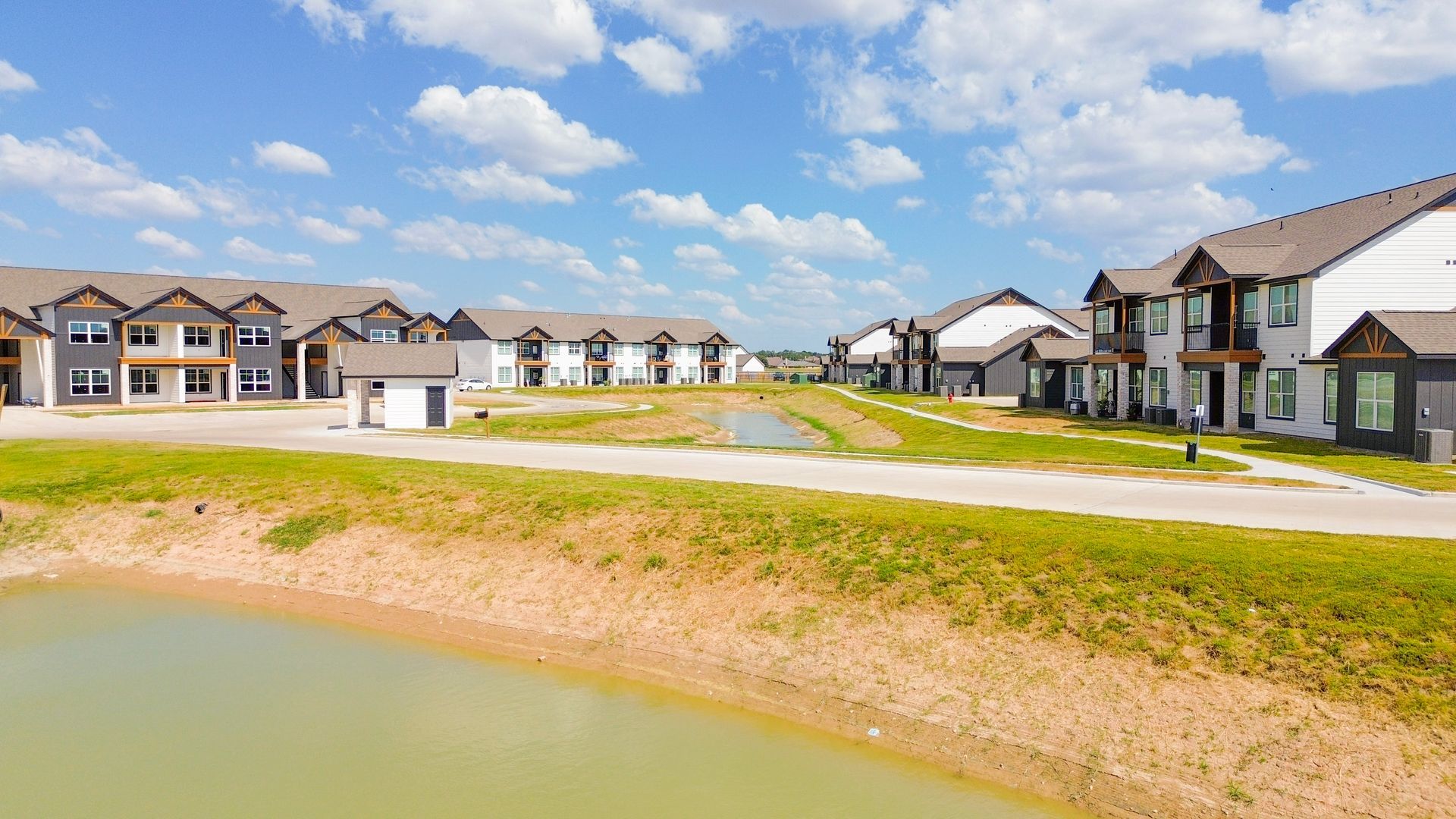 Rows of townhouses next to a pond under a bright blue sky with puffy clouds.