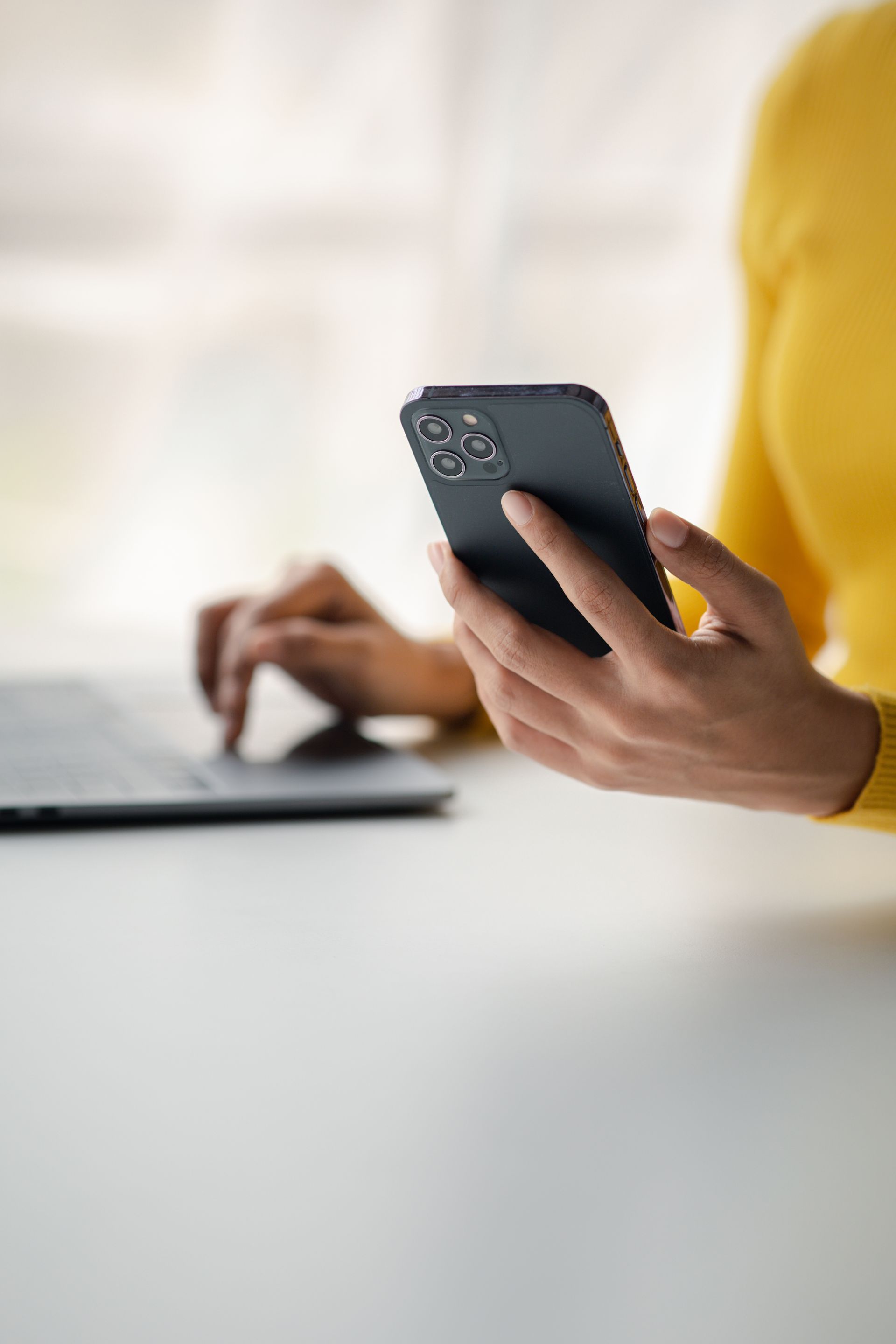 Person in yellow sweater holding a phone, typing on a laptop, close-up shot.