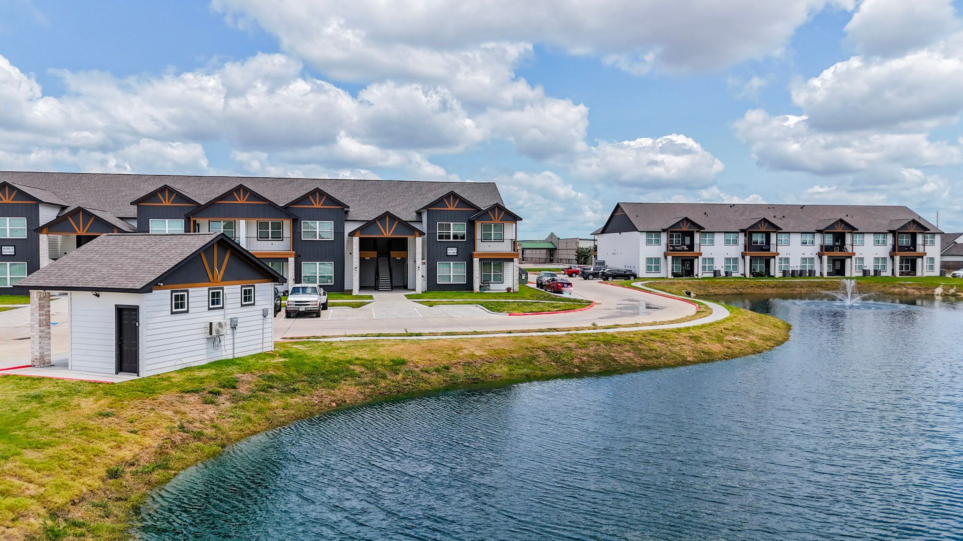 Aerial view of new apartment complex with brown roofs and a pond near a highway.