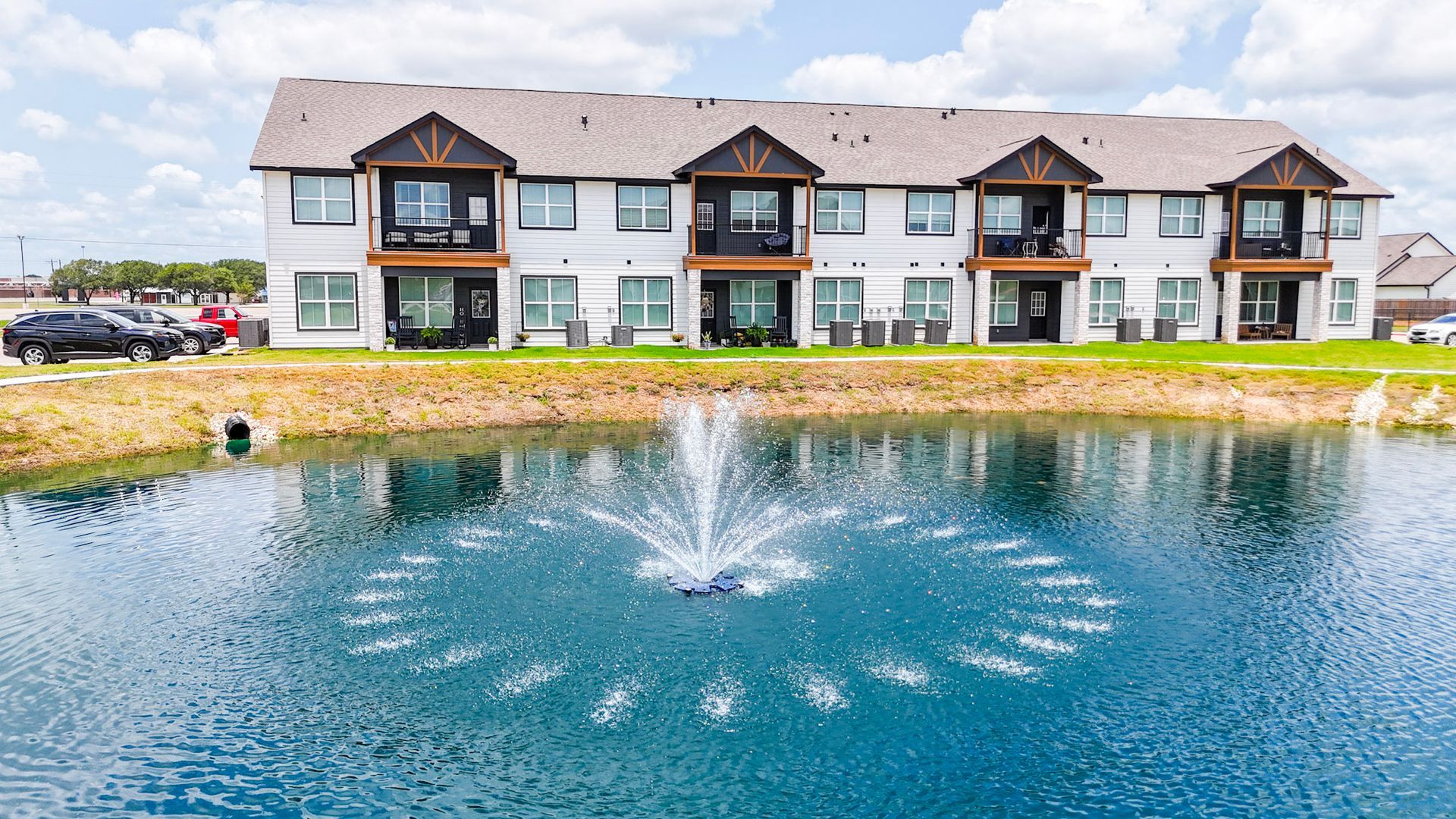 Exterior view of modern white apartments with brown roofs and parking lot, set in a rural area on a sunny day.