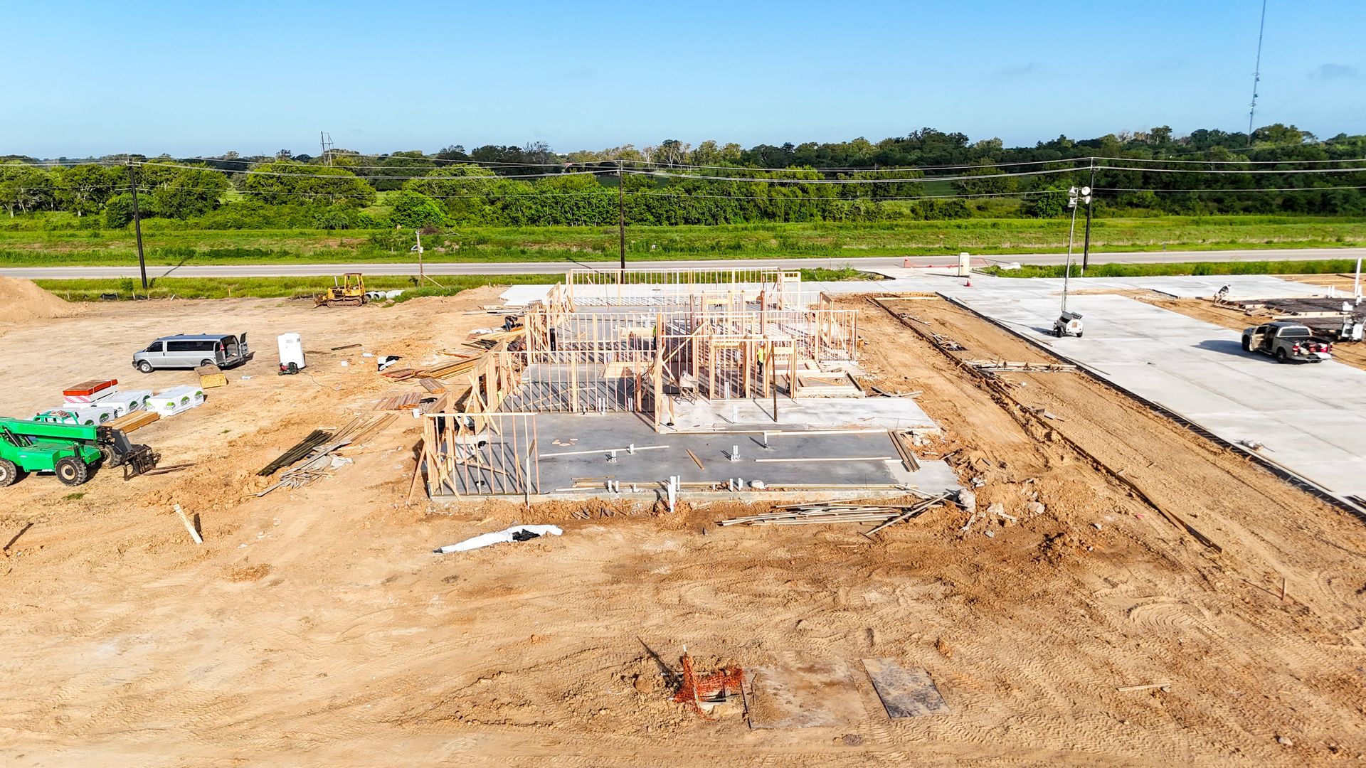 Construction site with wooden framing on a concrete foundation, surrounded by dirt and a paved area.