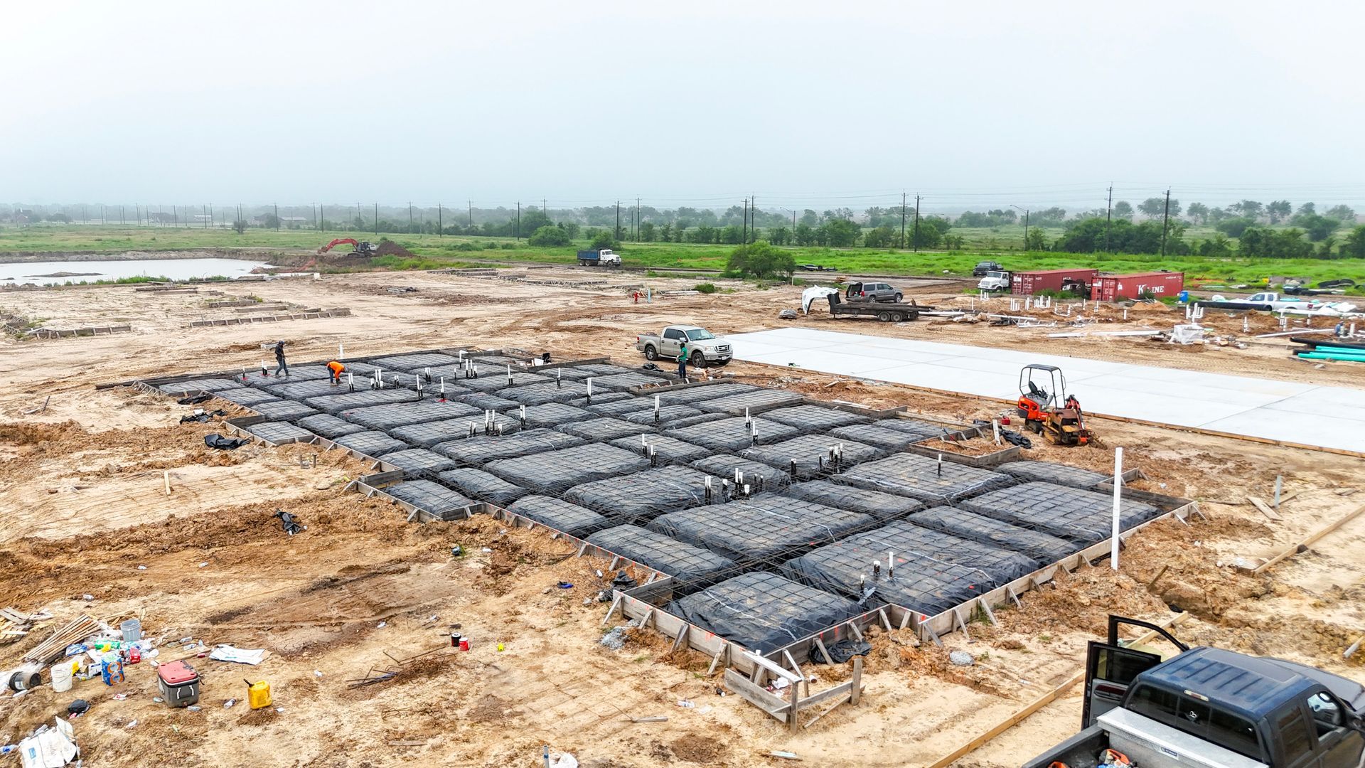 Construction site, workers building concrete foundation on leveled ground, with equipment and a road in the background.