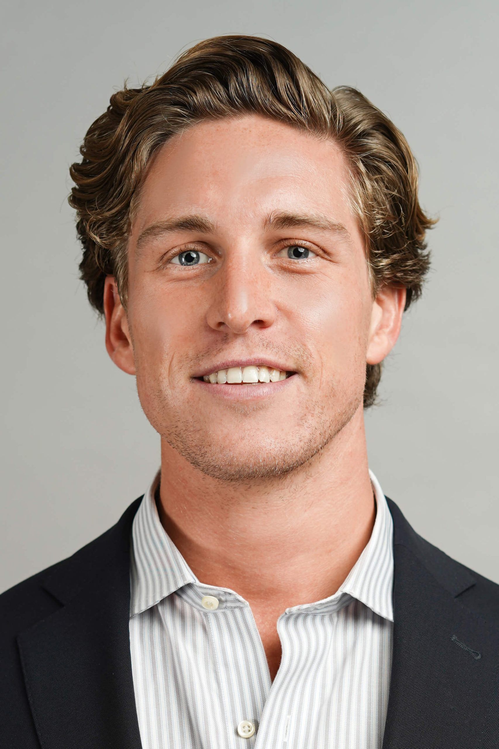 Man smiling, wearing a blue blazer and light blue button-up shirt against a neutral background.