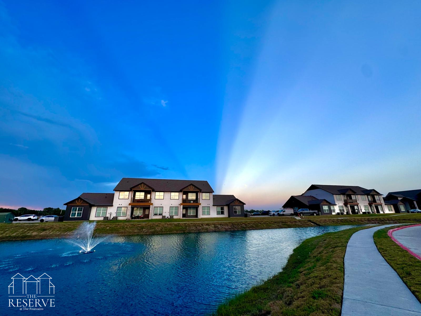 Apartment buildings near a pond at sunset, with blue and orange sky and sun rays.