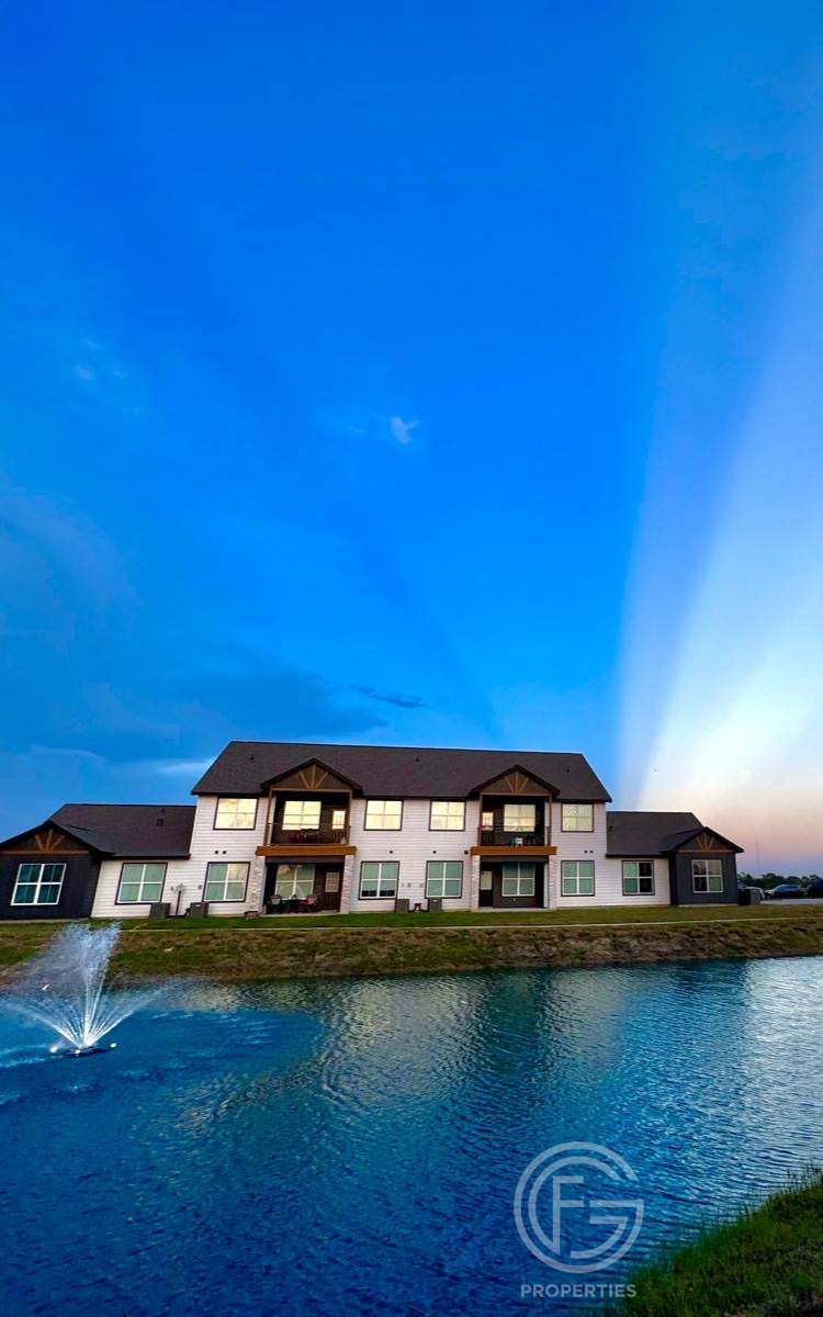 Modern two-story building with white exterior, brown trim, and a small lake. Dramatic blue sky with light beams.