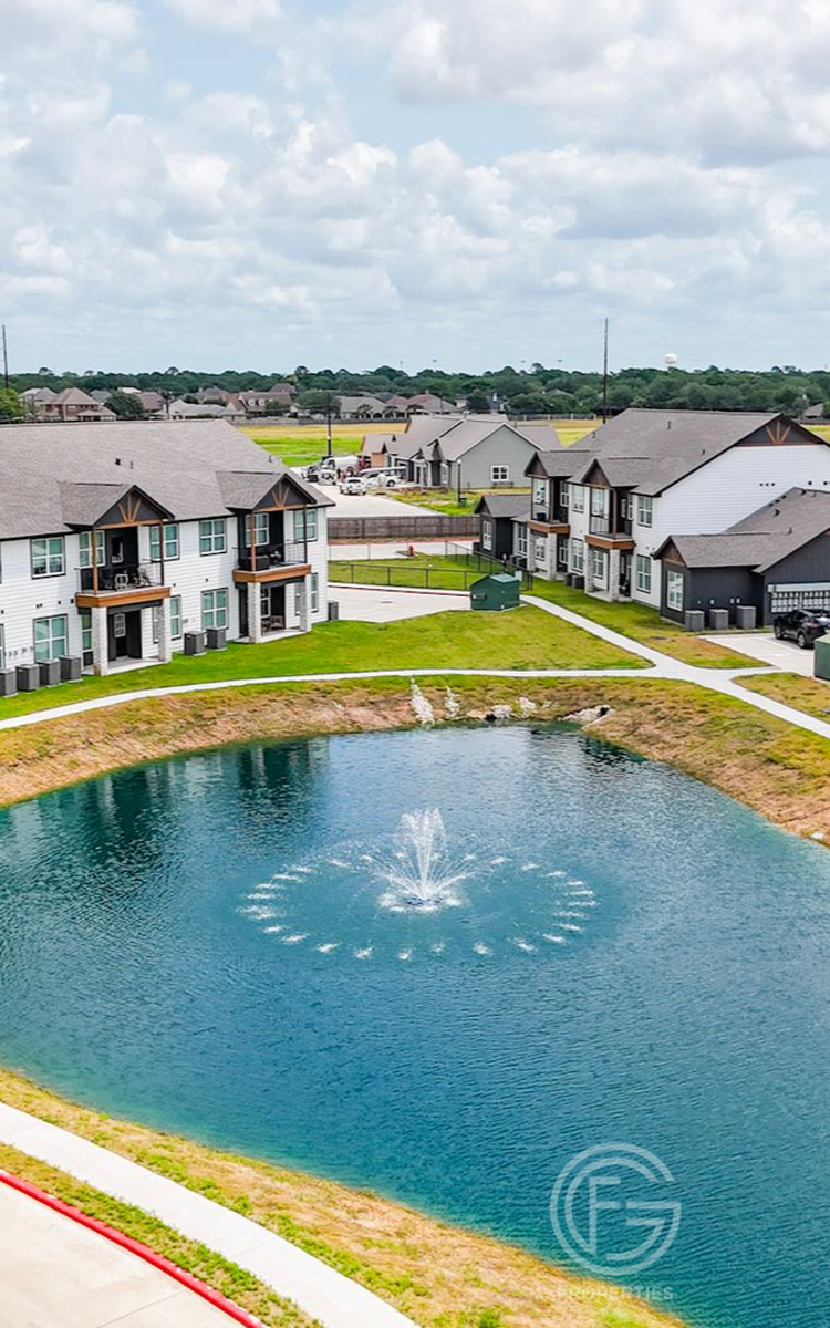 Apartment complex with pond featuring fountain, blue water, and green grassy areas on a sunny day.