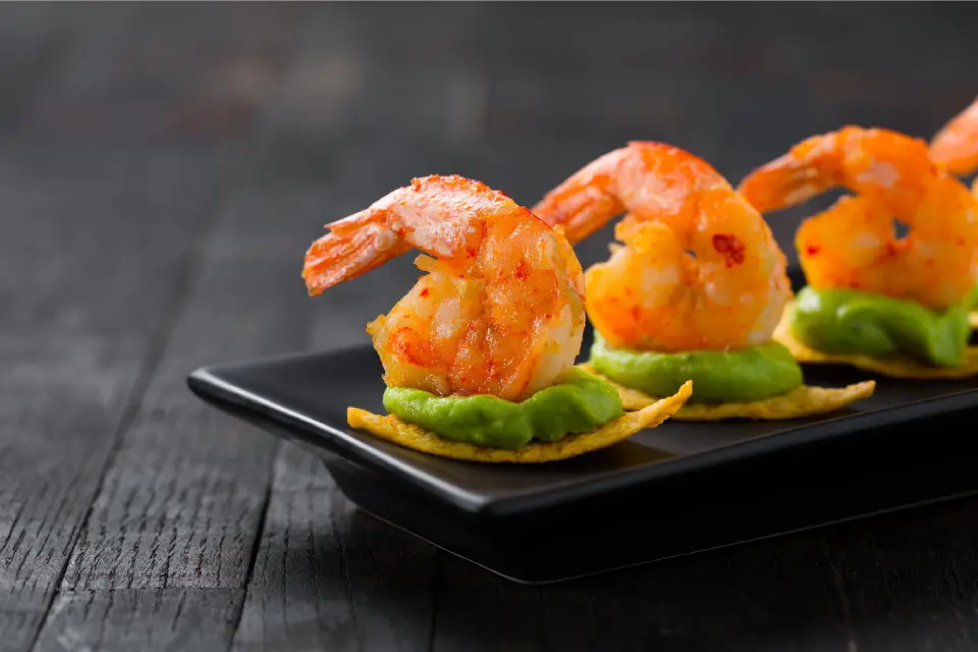 Une assiette noire garnie de crevettes et de guacamole sur une table en bois.