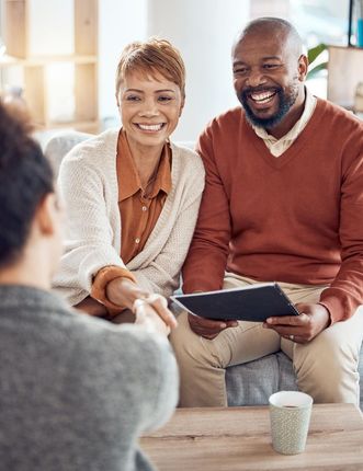 Two smiling people seated on a couch, holding a tablet and talking in a bright living room.