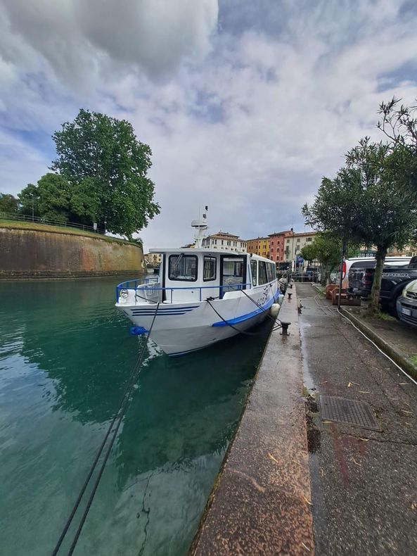 Barca bianca e blu ormeggiata su un canale accanto a un marciapiede bagnato e auto parcheggiate sotto un cielo nuvoloso