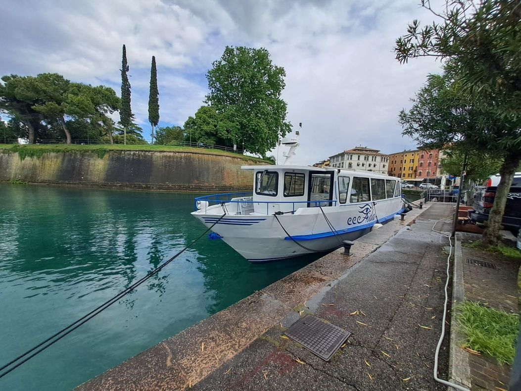 Una barca bianca ormeggiata su un fiume turchese, accanto a una passerella di pietra e ad alberi, sotto un cielo nuvoloso.