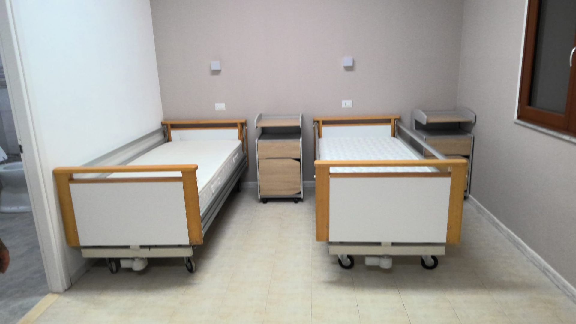Stanza d'ospedale con due letti vuoti, un comodino e pareti beige.