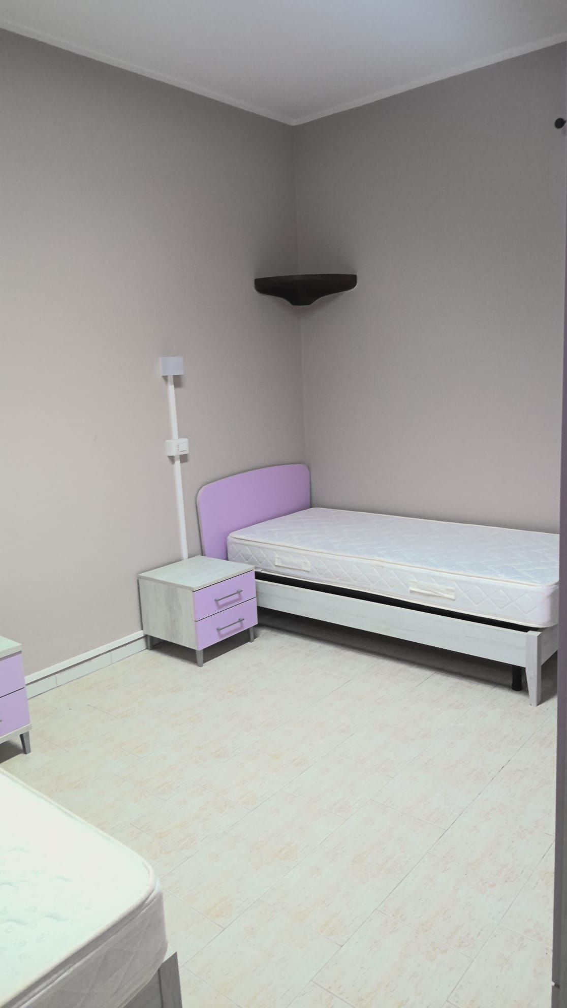 Piccola stanza d'ospedale con due letti singoli, pareti grigio chiaro e una lampada da comodino.