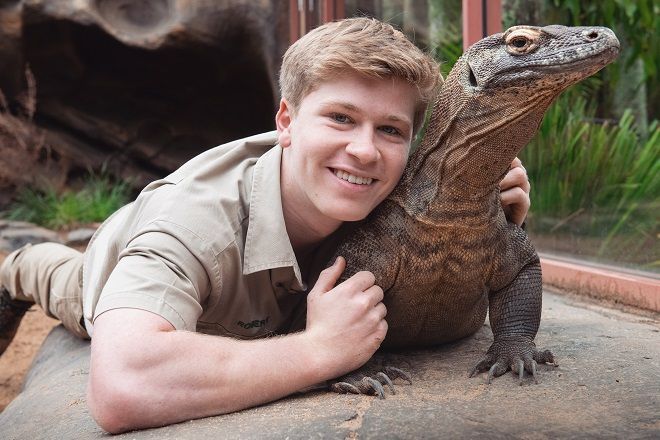 Man smiling, lying next to and hugging a Komodo dragon, on a rock outdoors.