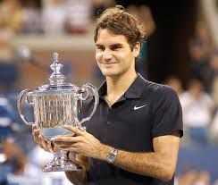 Roger Federer holding a trophy, smiling. He's in a black shirt with a watch; crowd in background.