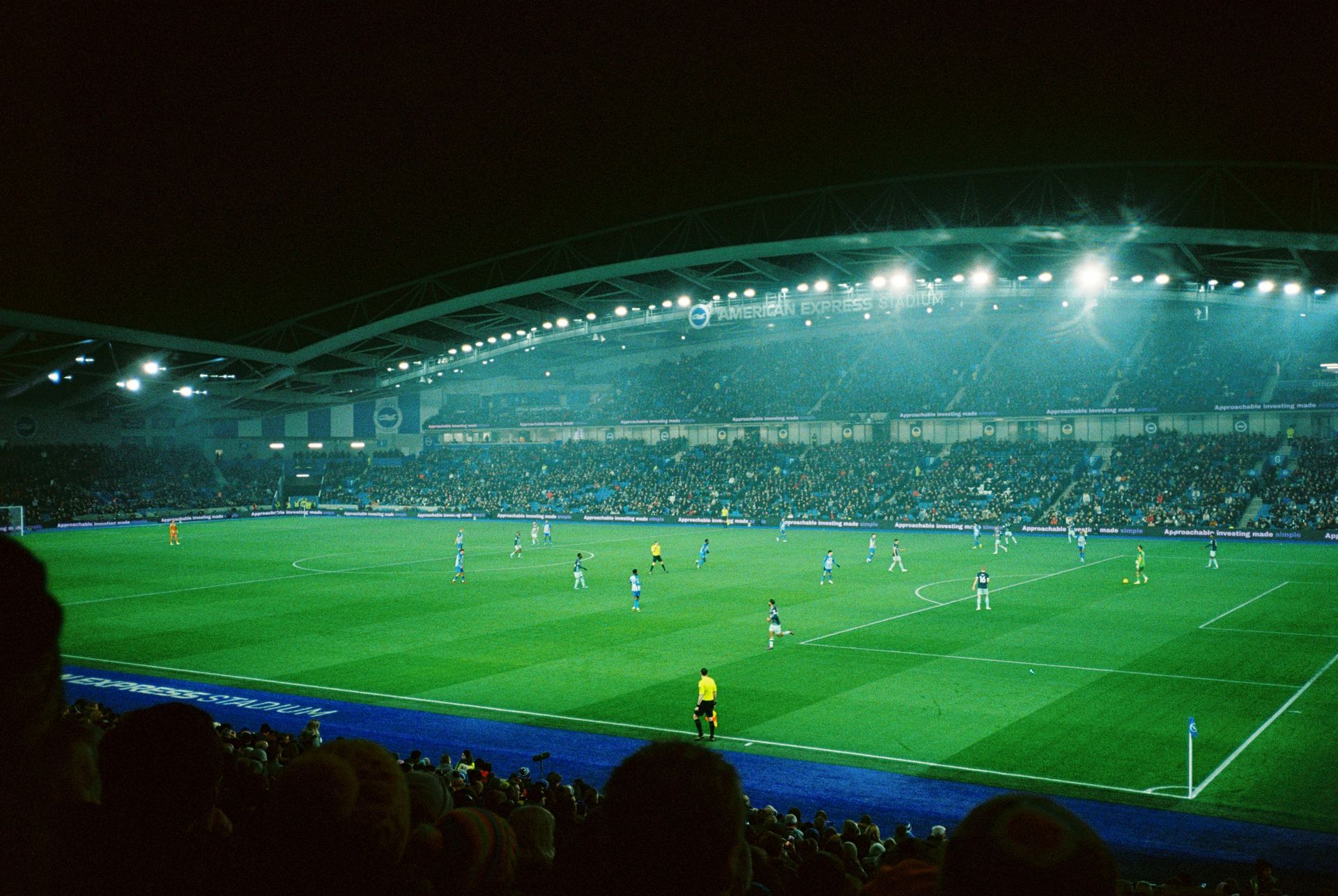 Soccer game at night in a stadium; green field with players, spectators in the stands, bright lights.