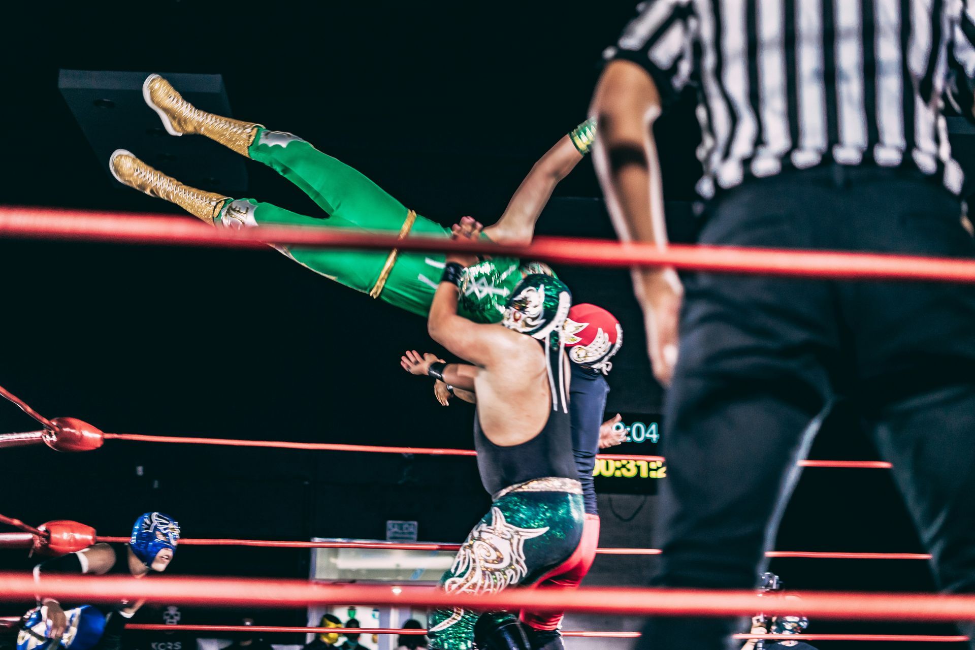 Wrestlers in colorful masks and outfits performing a high-flying maneuver in a wrestling ring, with a referee observing.