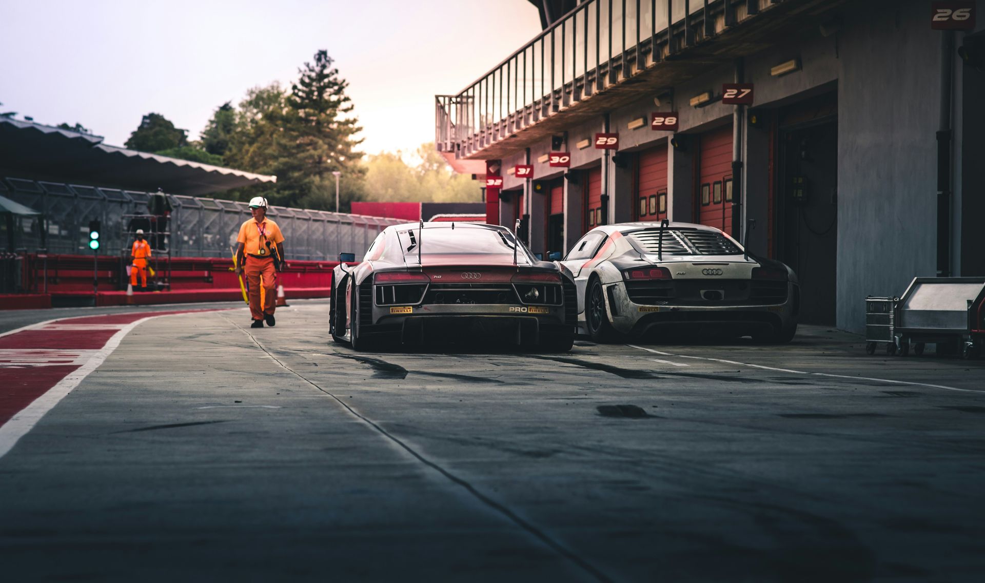Two race cars parked in front of garage doors, with a person in an orange suit walking nearby on a racetrack.