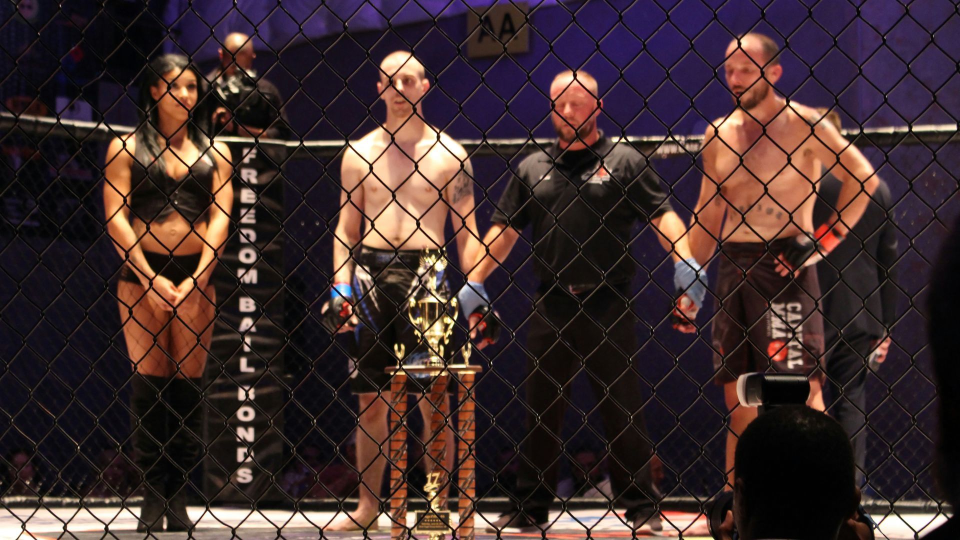 MMA fighters in cage with trophy, referee, and ring girl.