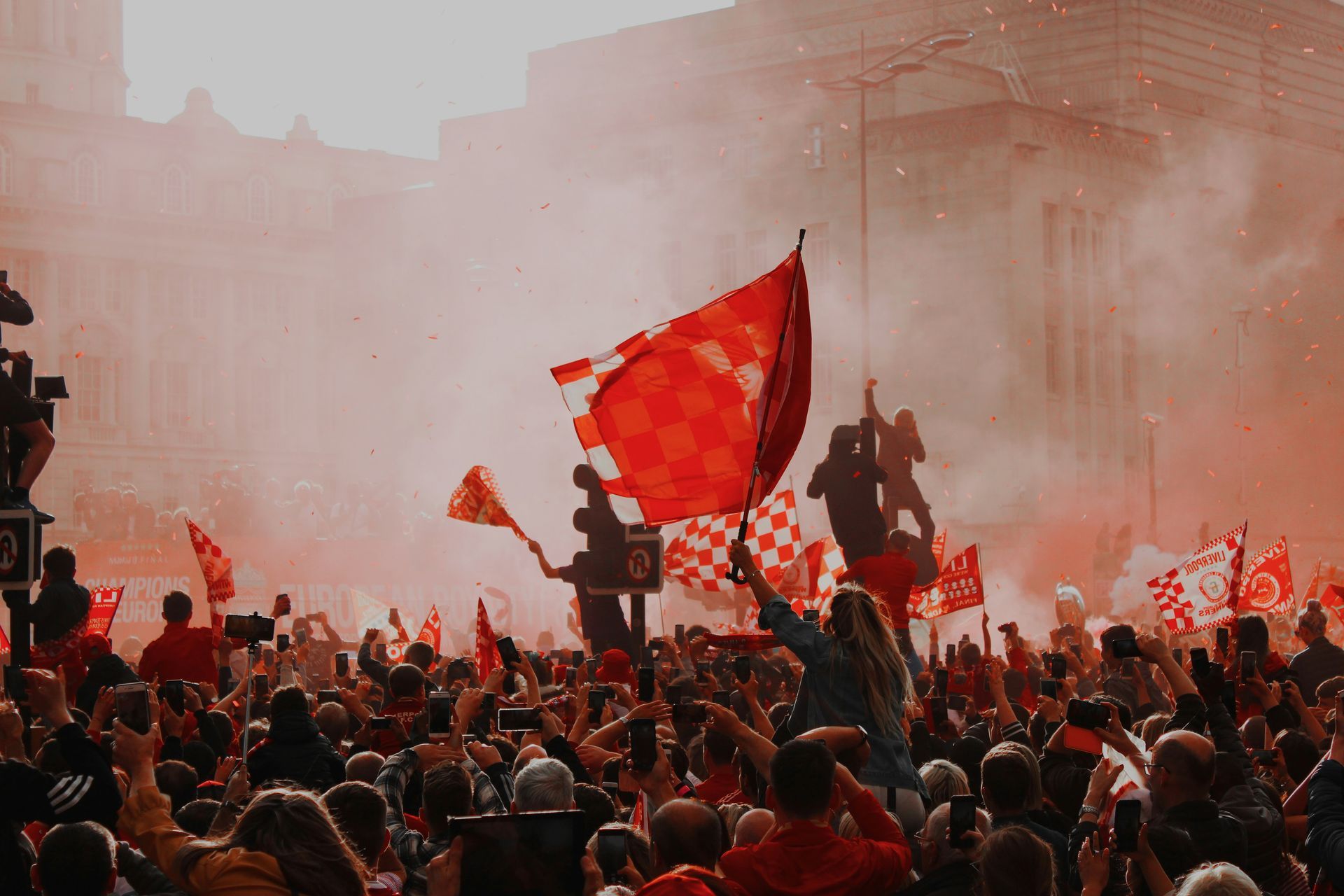 Crowd of people waving red flags in a city square, engulfed in red smoke and celebrating.