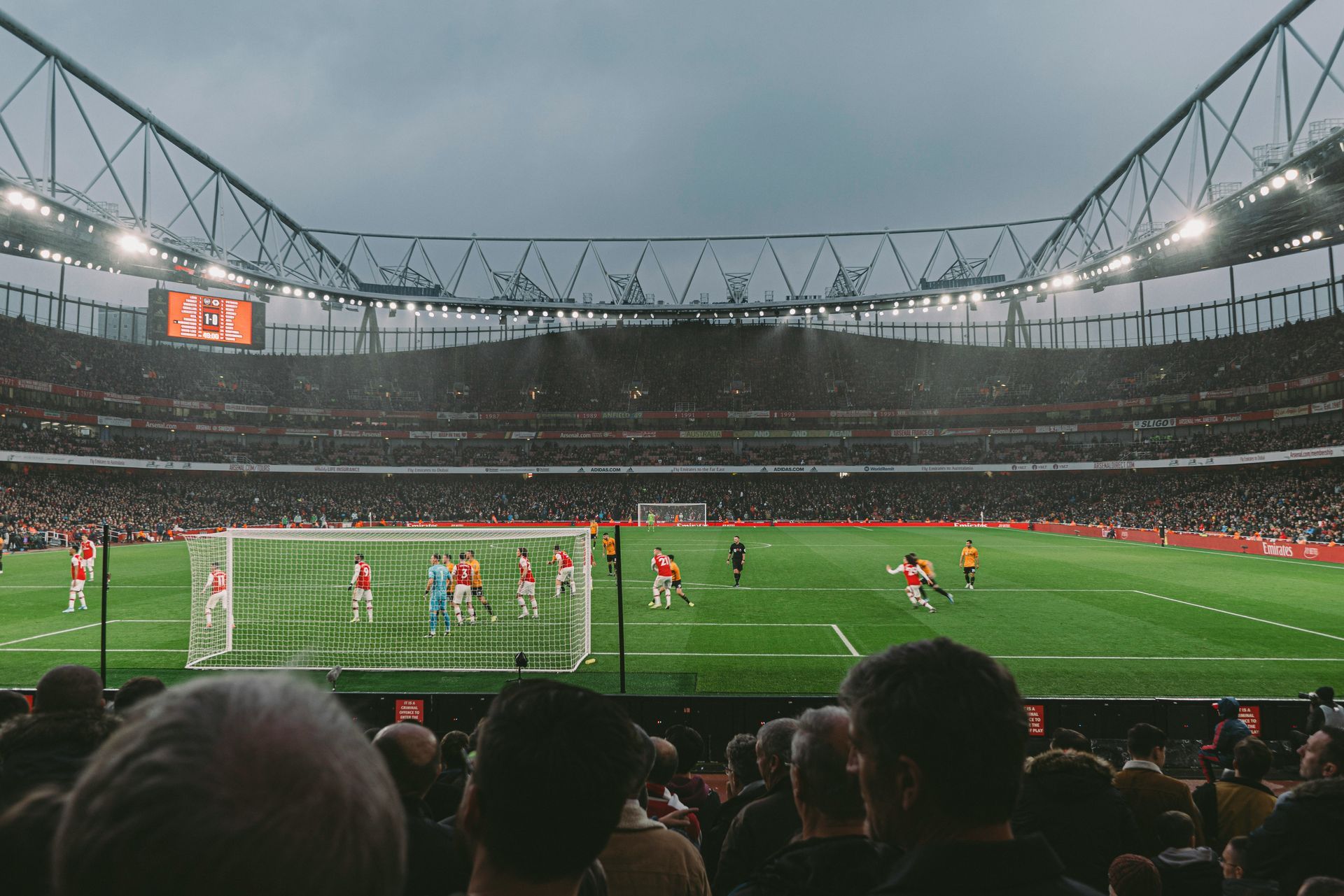 Soccer game in a packed stadium; players in red and white on the field, fans in the stands under a cloudy sky.