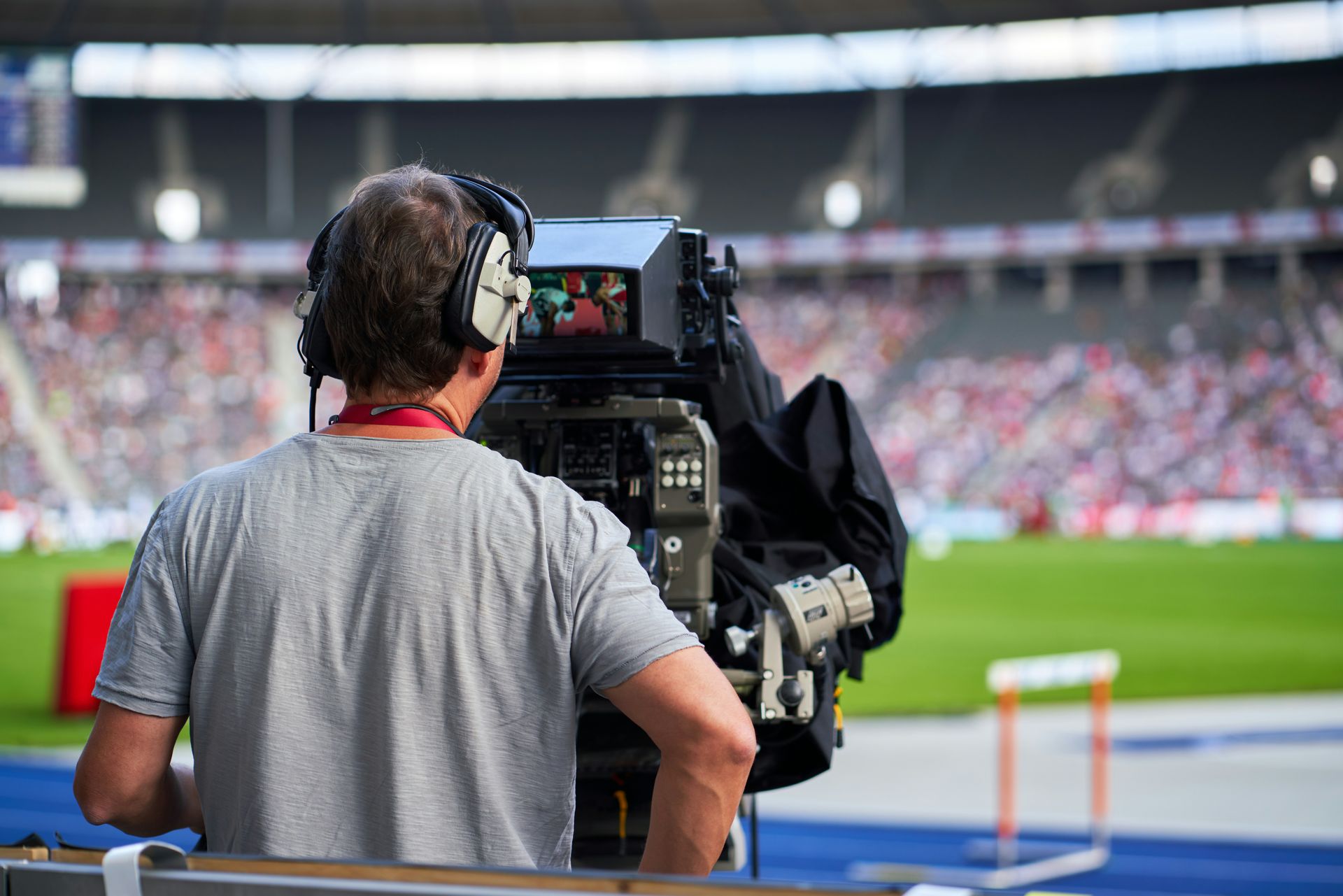 Camera operator filming a sporting event at a stadium. Focus on a camera, a blurred background with spectators, and a track.